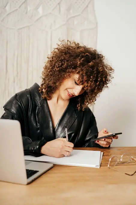 Positive retro businesswoman with curly hair smiling and writing in notebook while sitting at table and using smartphone in office