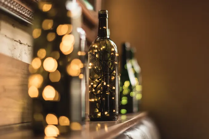 Composed transparent glass bottles with sparkling light garlands inside placed in row on wooden shelf near wall