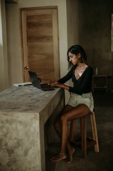 Serious focused youthful ethnic female typing on laptop while sitting at table on table kitchen modern marble countertop