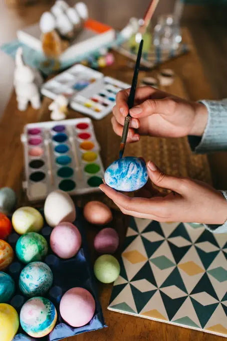 Unrecognizable person in sweater covering fresh chicken eggs with blue paint while preparing for Easter celebration