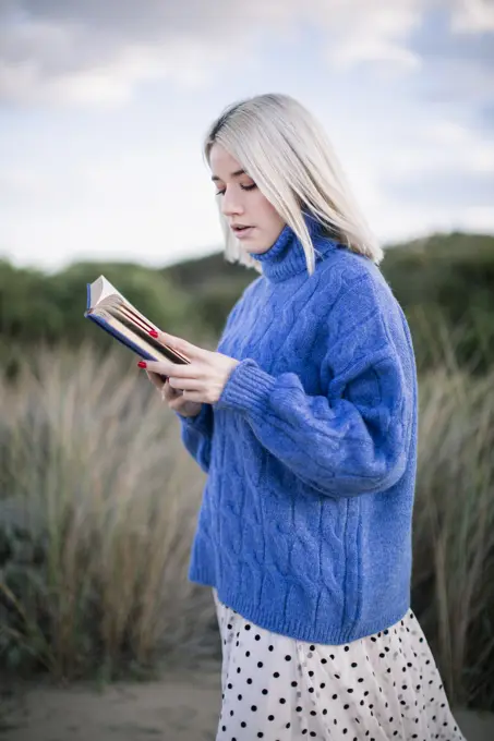 Thoughtful side view of young woman with blonde hair in warm blue sweater holding open book reading while standing against blurred natural sandy background