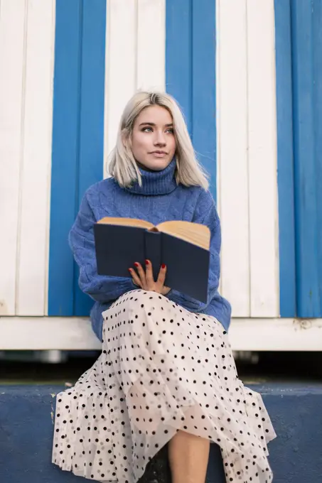 From below thoughtful young female in warm sweater and skirt sitting with open book reading against white and blue striped wall looking away