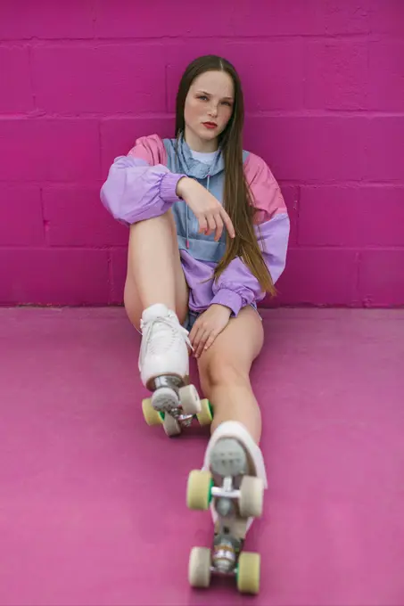 Serious confident female teenager in stylish colorful sports jacket and quad roller skates looking at camera while sitting against pink background