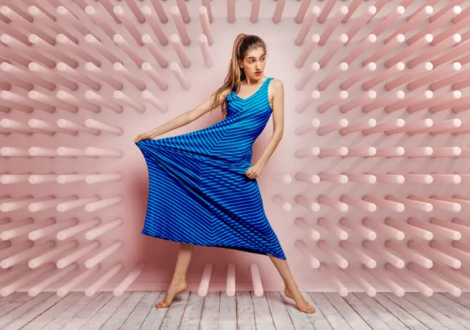 Young woman barefoot looking away stretching side of a blue dress while standing on tube hook pink background