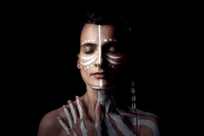 Portrait of naked tender beautiful brunette Native American woman with white striped painted on body covering breast standing in dark on black background with closed eyes