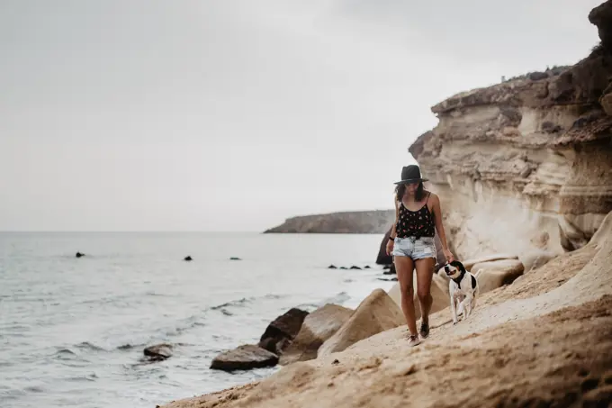 Trendy young lady in black hat and casual clothes walking along rocky coast with obedient dappled dog against troubled sea water under gray cloudy sky