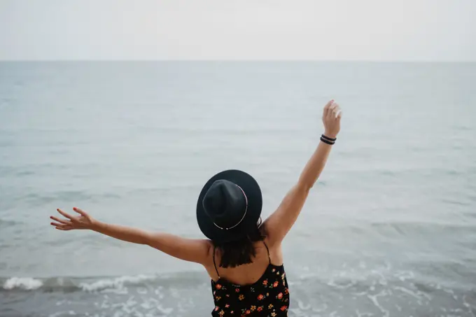 High angle back view of female in black hat and dress standing with arms raised and enjoying life on beach against troubled ocean waves in overcast weather