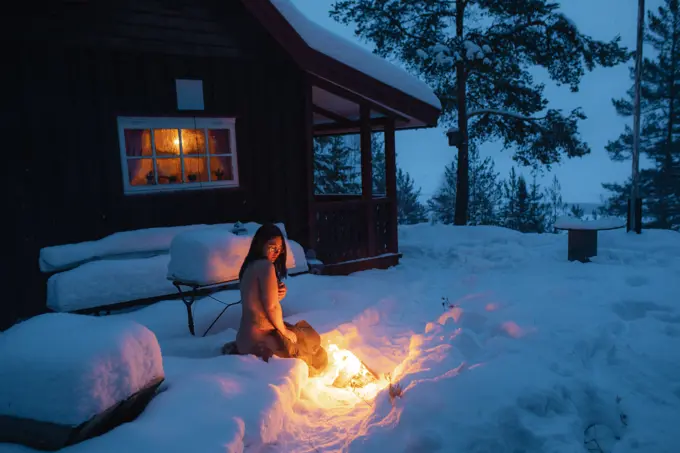 Side view of young nude woman with tattoos sitting in snow against rural house looking at bonfire near