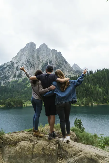 Back view of two women and man embracing and having fun at lake in mountains together.