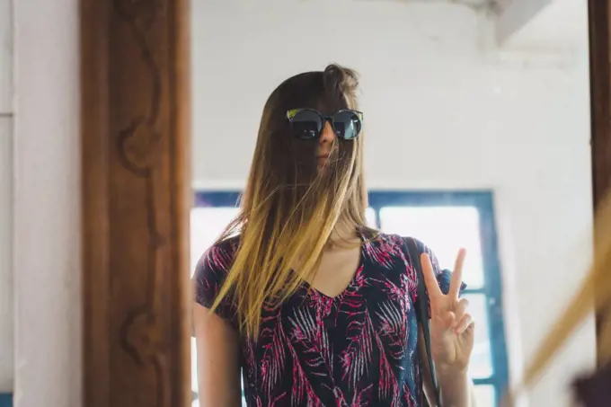 Pretty woman standing at mirror with sunglasses over hair and gesturing two fingers.