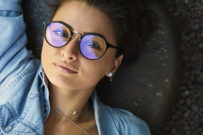 Top view of young stylish girl wearing elegant eyeglasses and looking at camera while lying on ground.