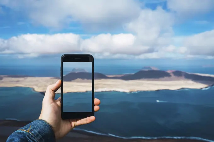 Unrecognizable tourist taking shot of small island in blue sea with smartphoneinLanzarote, Spain.