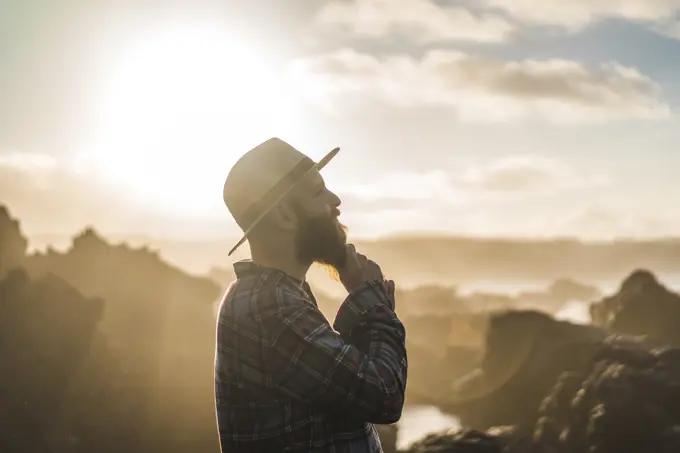 Side view of unrecognizable tourist in hat standing on cliff in sunset lights.