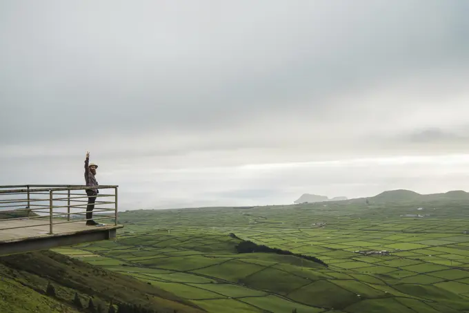 Side view of tourist standing on viewpoint with hand up on background of green fields.