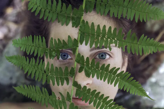 From above shot of girl looking at camera while covering face with green fern leaf.