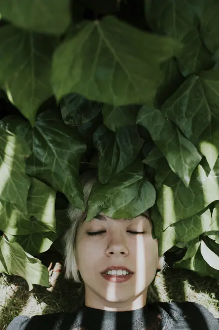 Portrait of young smiling girl with blonde hair resting with eyes closed while lying on green leaves in sunlight.From above