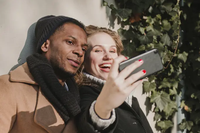 Happy multiethnic couple in warm clothes standing at bush and taking selfie.