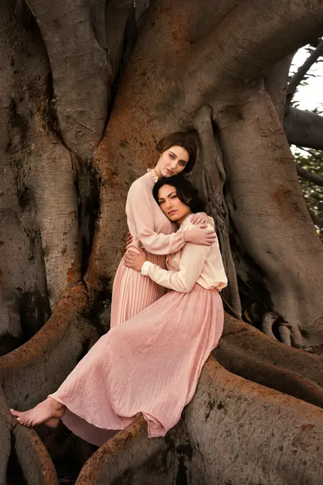 Pretty women in vintage dresses looking at camera and embracing at huge tree.