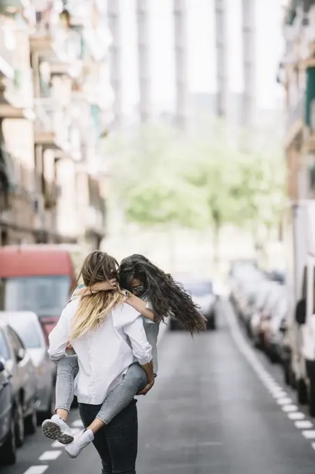 Back view of young woman carrying her girlfriend near cars on road. Vertical outdoors shot. 