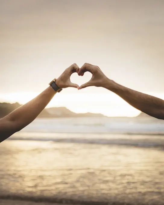 Two unrecognizable men gesturing heart with hands in sunset lights. 