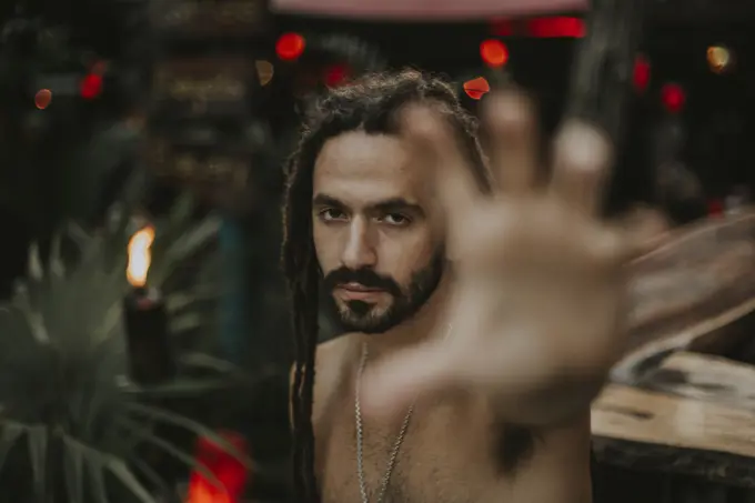 Portrait of man with dreadlocks posing without shirt outstretching hand at camera on background of tropics.