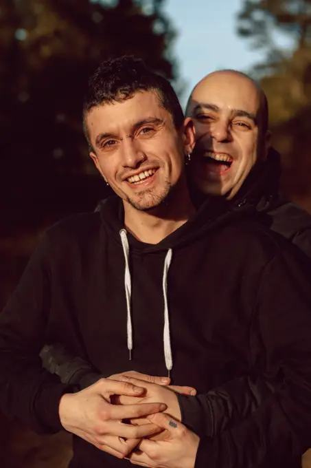 Cheerful homosexual couple embracing on walkway in forest in sunny day on blurred background