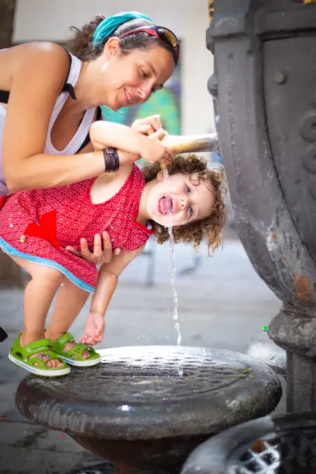 Happy mother pouring water from drinking fountain for funny girl with opened mouth on street in summer