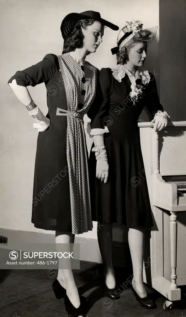 Two young women in dresses and hats posing by piano