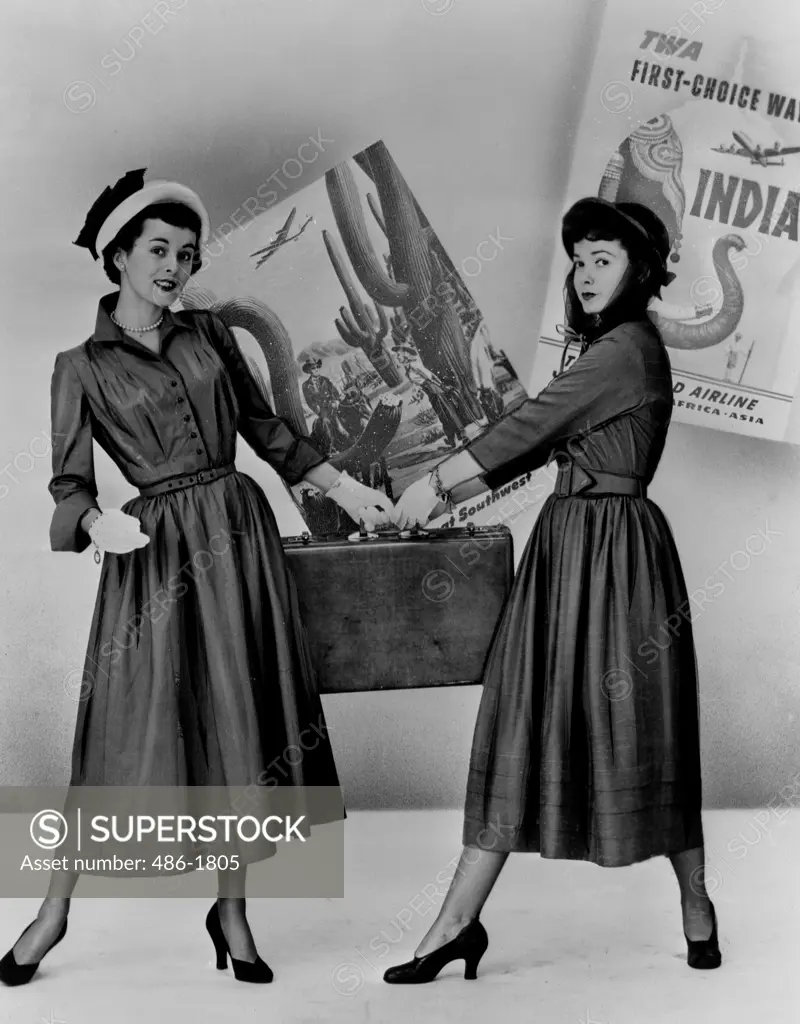 Two fashion models posing with suitcase, posters in background