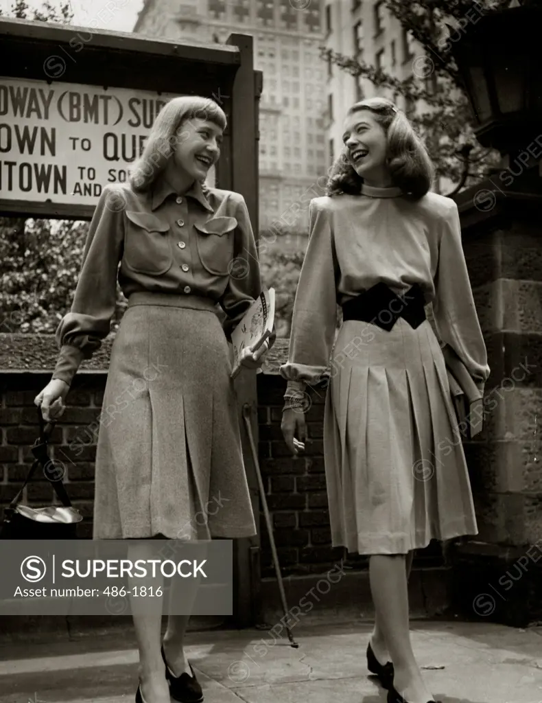 Two young women walking on street in elegant clothing, smiling face to face