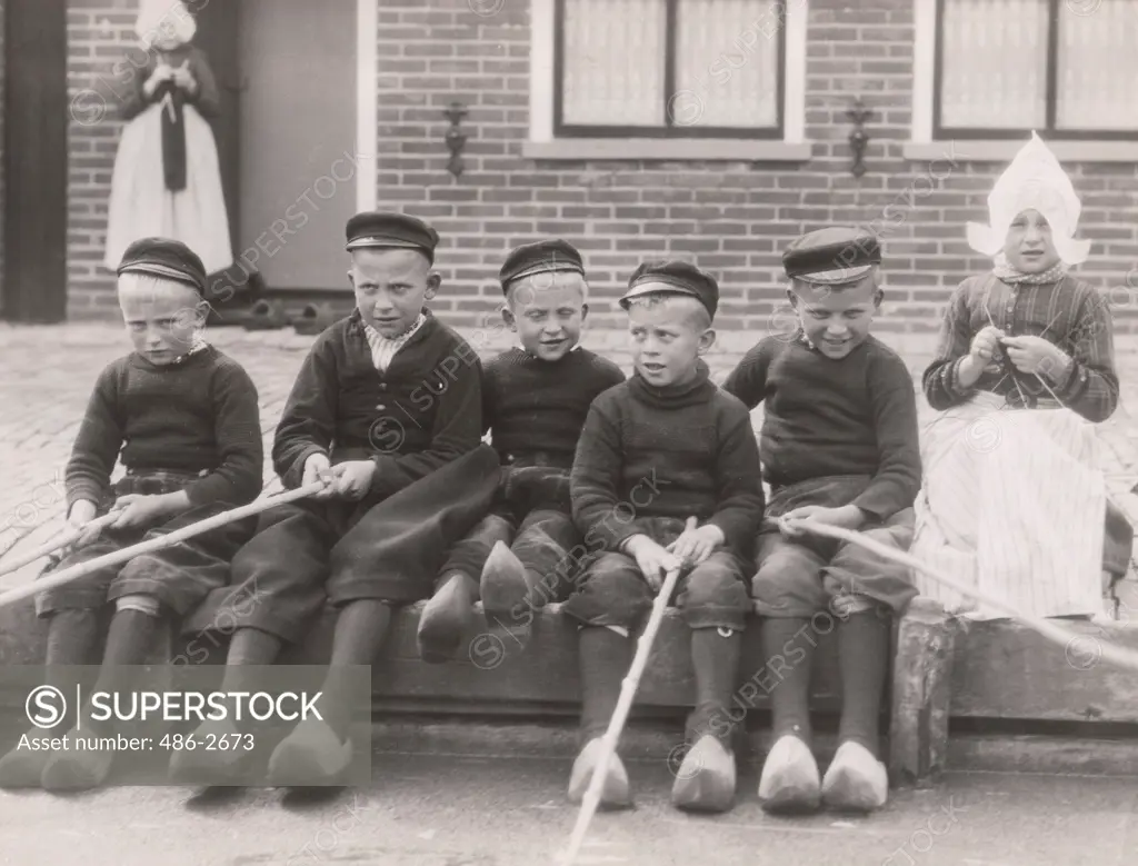 Boys and girl sitting on bench in front of building, holding sticks