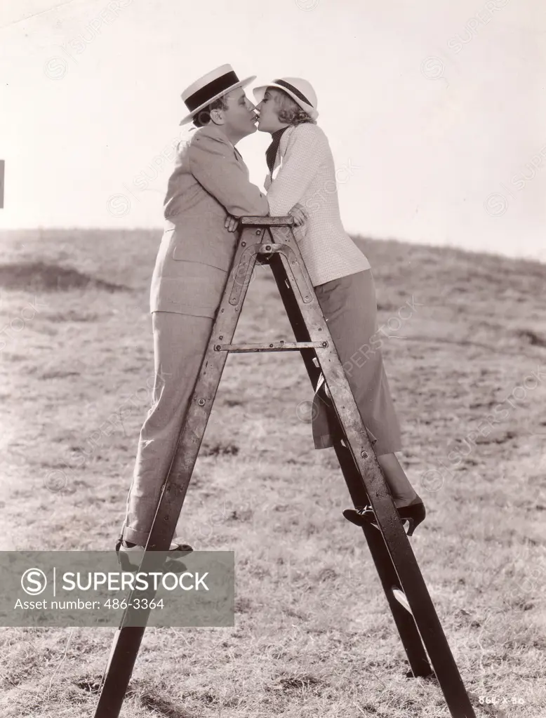 Scene from movie ""Let Freedom Ring"" directed by Charles F. Riesner with Une Merkel and Jack Benny kissing on top of ladder, Metro-Goldwyn-Mayer production, Kissing up ladder to love. Une Merkel and Jack Benny find top of ladder excellent place in which to practice one of love scenes in their latest Metro-Goldwyn-Mayer production, ""Let Freedom Ring"", which Charles F. Riesner is directing