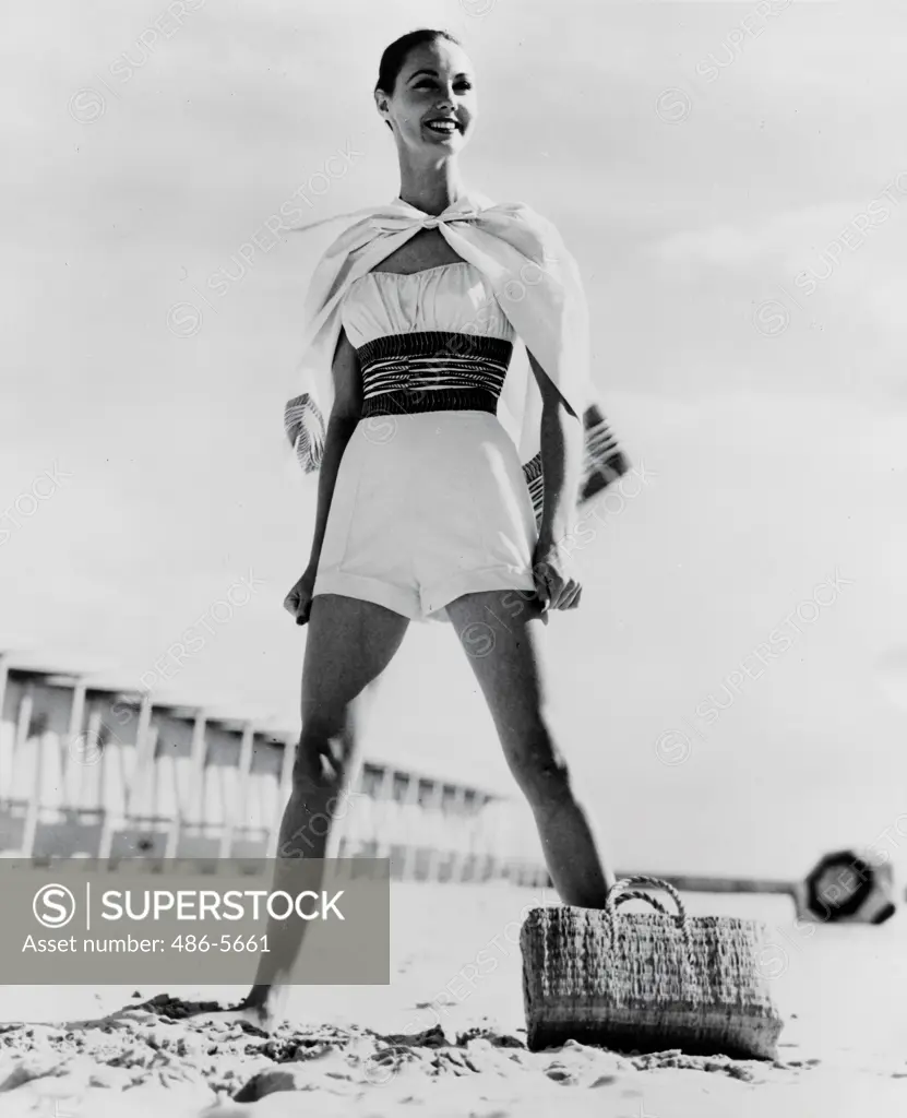 Fashion shoot of one piece swimwear, 1957, Schiaparelli Accent the ""Little boylook"" in a one-piece swimsuit of white cotton. A wide print cummerbund nips in the waist and there's a matching cape (also worn as a skirt) with a colorful trim to match the cummerbund, 1957