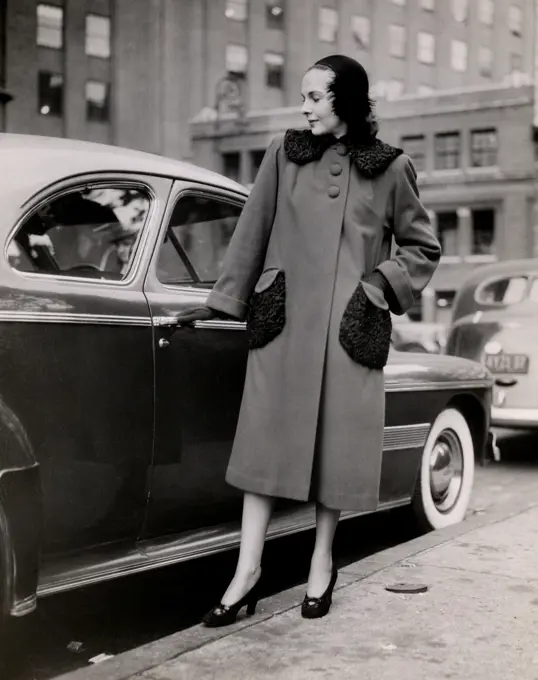 Mid adult woman wearing coat, posing in front of parked car, 1931