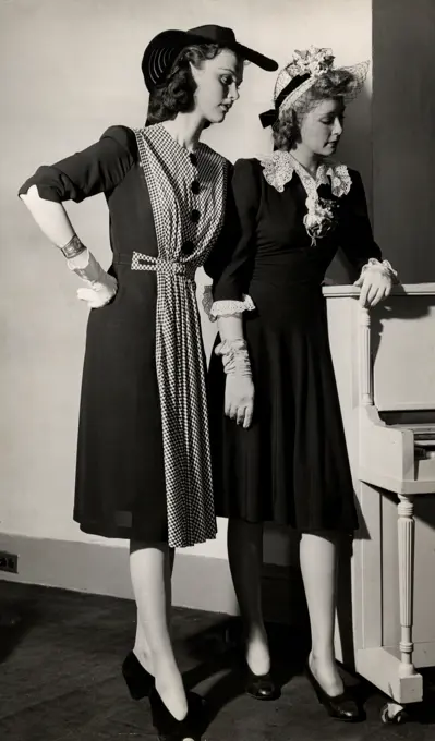 Two young women in dresses and hats posing by piano