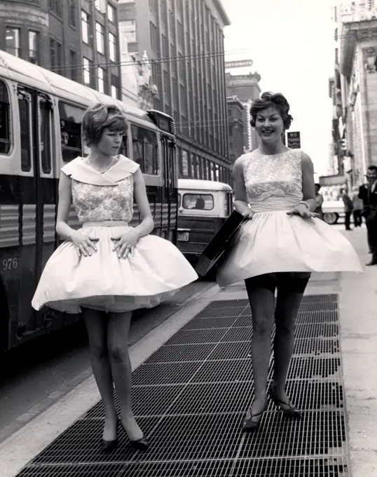 Two young women in short dresses and stockings walking over sidewalk vent