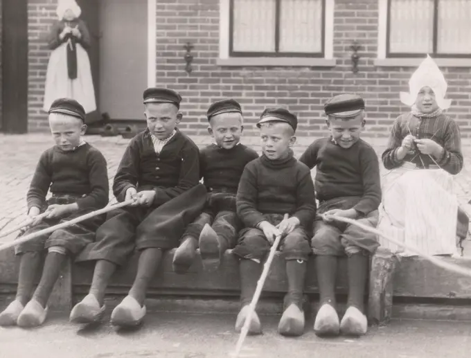 Boys and girl sitting on bench in front of building, holding sticks