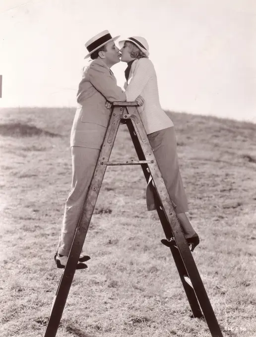 Scene from movie ""Let Freedom Ring"" directed by Charles F. Riesner with Une Merkel and Jack Benny kissing on top of ladder, Metro-Goldwyn-Mayer production, Kissing up ladder to love. Une Merkel and Jack Benny find top of ladder excellent place in which to practice one of love scenes in their latest Metro-Goldwyn-Mayer production, ""Let Freedom Ring"", which Charles F. Riesner is directing