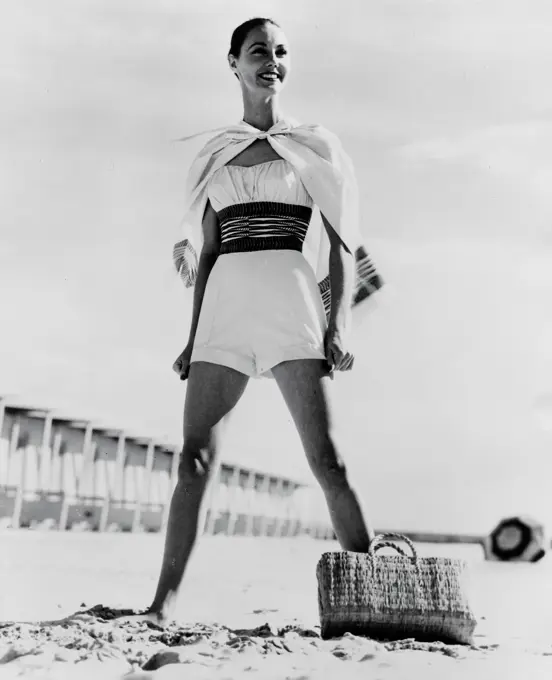 Fashion shoot of one piece swimwear, 1957, Schiaparelli Accent the ""Little boylook"" in a one-piece swimsuit of white cotton. A wide print cummerbund nips in the waist and there's a matching cape (also worn as a skirt) with a colorful trim to match the cummerbund, 1957