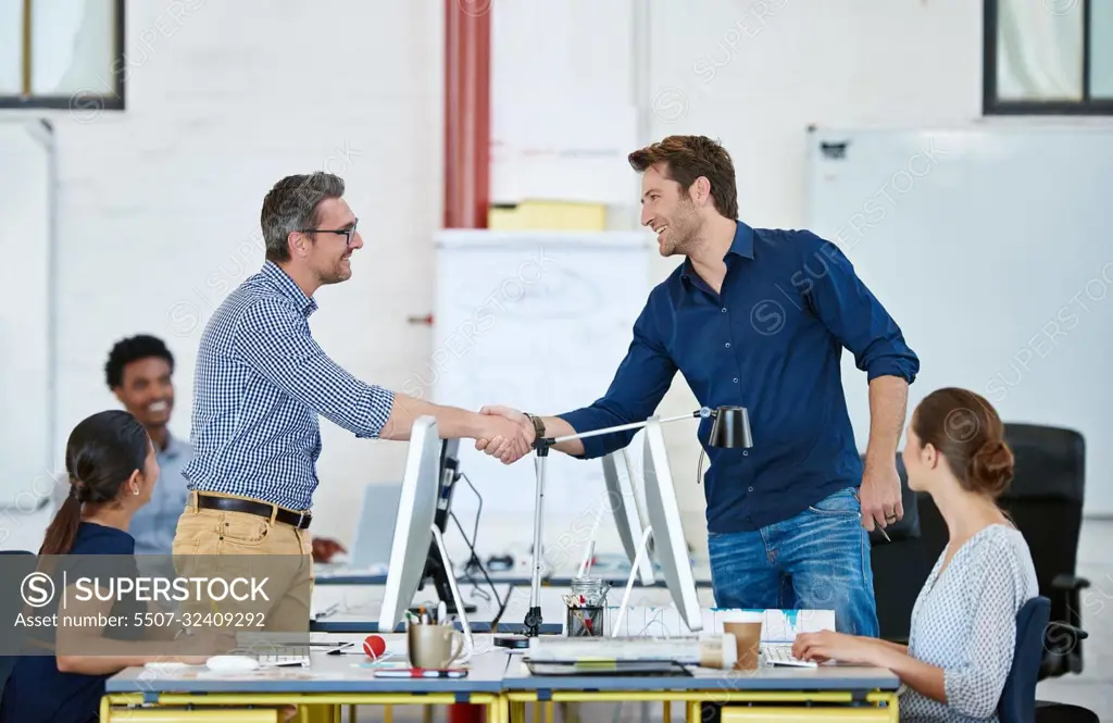 Another job well done. two designers shaking hands over their computers in an office.