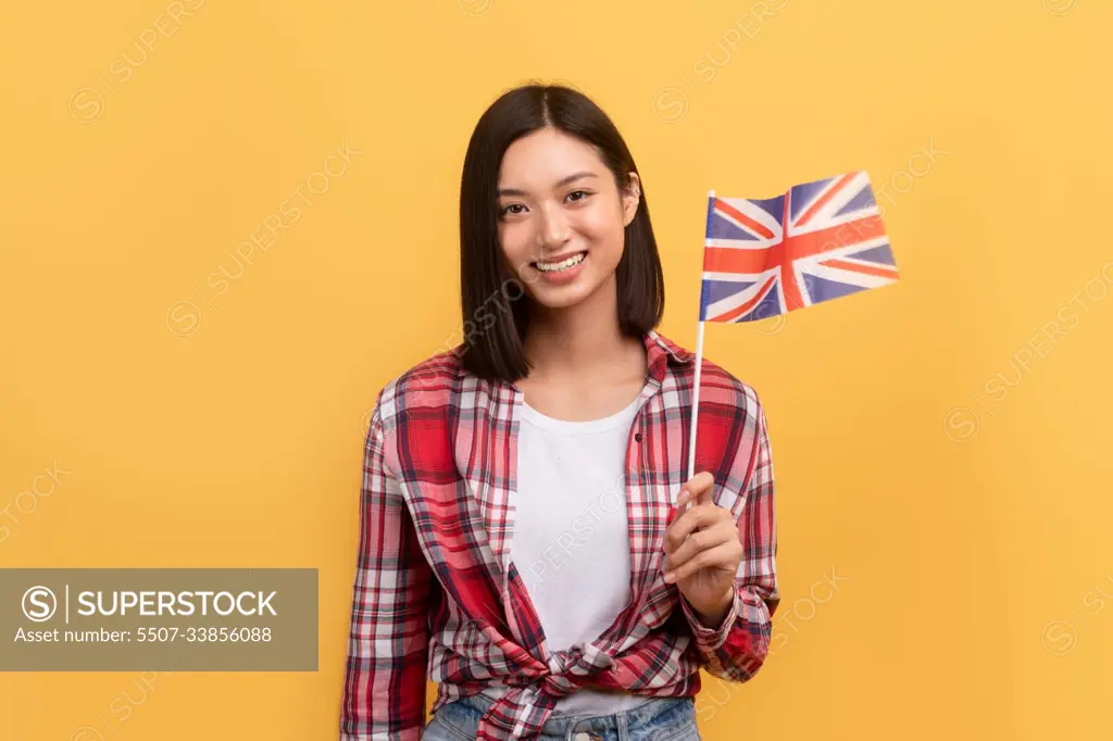 Cheerful asian lady student holding England flag, enjoying tutoring or studying language, posing on yellow background