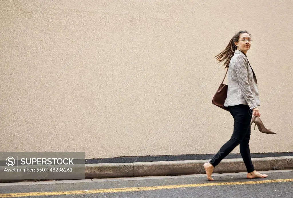 Woman in street, walking at wall with mockup and barefoot, relax and travel for work holiday in city. Space, road and freedom, person on walk with shoes in hands, morning commute and business trip.