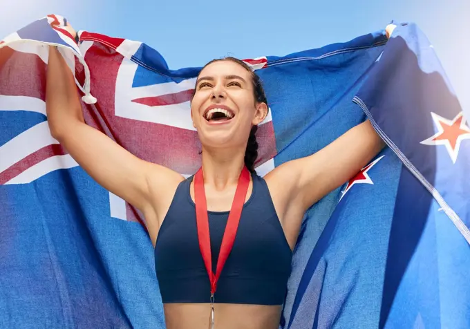Young fit female athlete cheering and holding a New Zealand flag with a gold medal after competing in the Olympics. Smiling active sporty woman feeling motivated and celebrating winning in sport