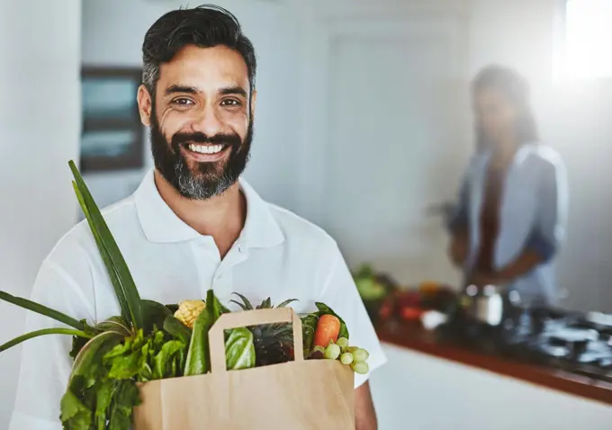 Buy healthy, eat healthy. Portrait of a happy man holding a bag of fresh vegetables while standing in his kitchen.