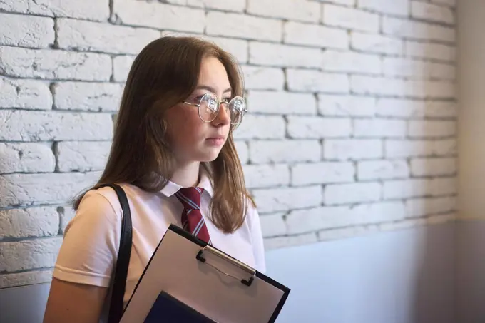 Portrait of university student girl, confident female posing in white T-shirt, glasses, tie, backpack. Background white brick wall, copy space