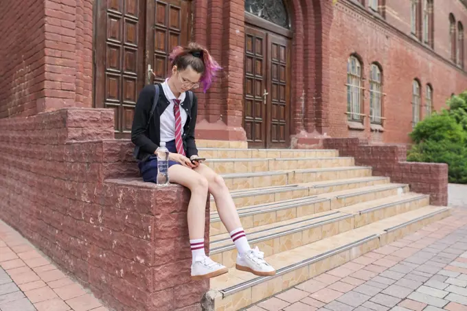 Schoolgirl teenager in uniform with backpack using smartphone