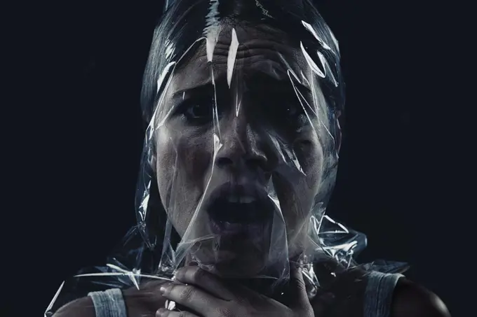 Fighting for oxygen, fighting for her life. A young woman suffocating with her head wrapped in plastic while isolated on a black background.