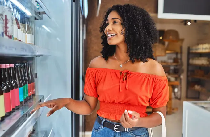 Happy, grocery shopping and woman in a supermarket with drinks at a retail store in Sao Paulo. Happiness, smile and girl from Brazil buying a beverage on a shelf in a food shop while on vacation.