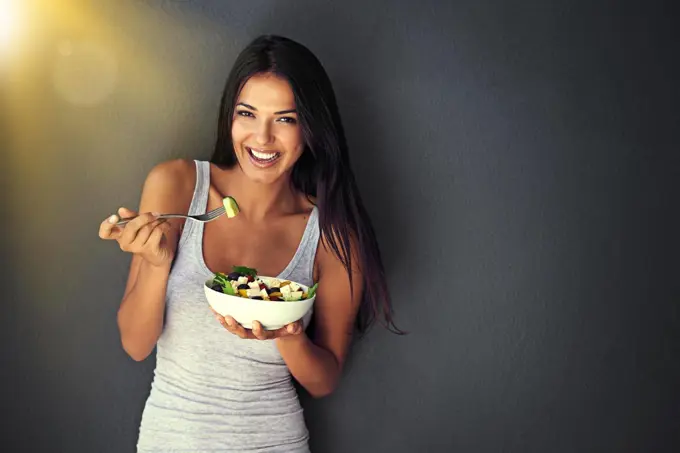 Healthy and delicious. Portrait of a healthy young woman eating a salad against a gray background.