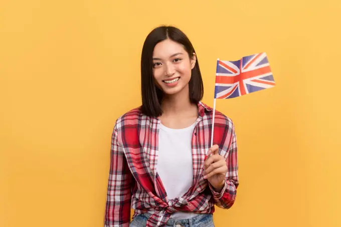 Cheerful asian lady student holding England flag, enjoying tutoring or studying language, posing on yellow background